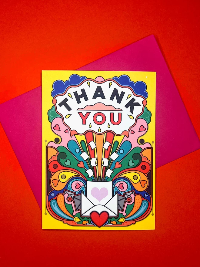 A vibrant yellow card with Thank You written boldly at the top, above a multi-coloured abstract design featuring hearts, envelopes and paper sits on a pink envelope on top of a red backdrop. 