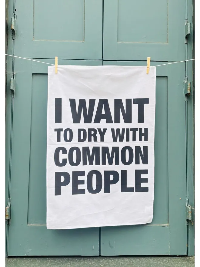 I Want to Dry with Common People screen printed tea towel hanging on washing line in front of teal shutters