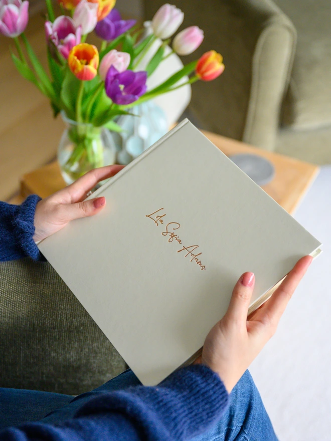 a woman is holding a pearly white photo album in her hands. The photo album has been printed on the front with a girl's name in a calligraphy writing. There is a big bunch of tulips in the background and the room feels bright and spring-like 