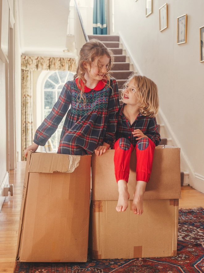 A boy and girl in matching navy tartan outfits hide in boxes