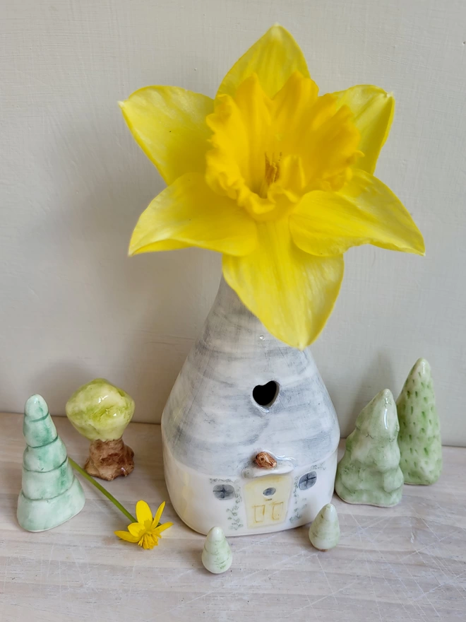 a group of ceramic hand painted trees surrounding a ceramic vase in the shape of a house with a yellow daffordil