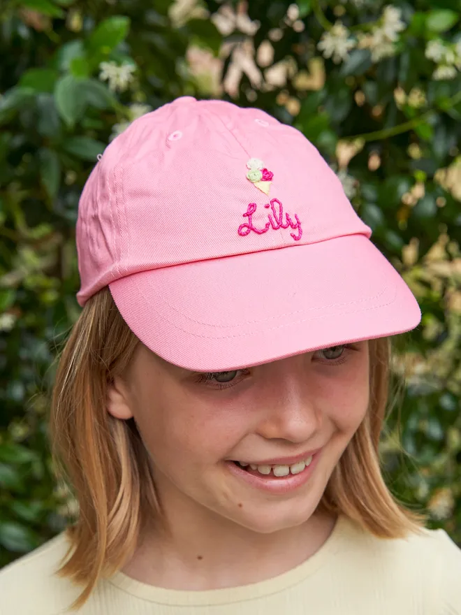Hand embroidered personalised children's cap in pink. The pink cap is worn by a girl and the cap is embroidered with flowers. 