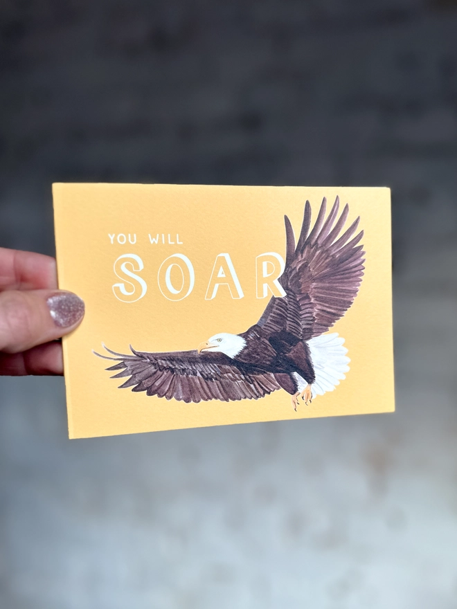 a bright yellow greetings card featuring an illustration of a bald eagle with the text “you will soar”