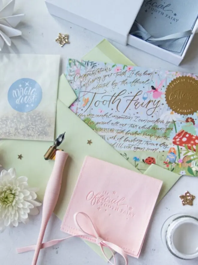 An illustrated garden notecard with fairies, toadstools and gold foil "Tooth Fairy" lettering rests on a pale green envelope. Beside it are a blush pink pouch, a Magic Dust packet and a pink calligraphy pen.