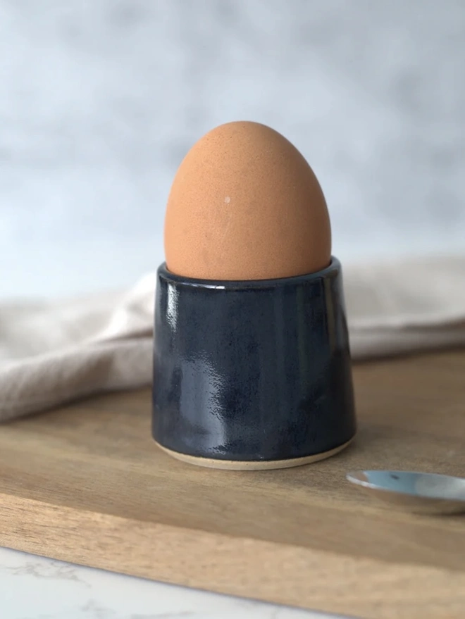 Classic Handmade Egg Cup, a ceramic egg cup sitting on a wooden chopping board against a grey backdrop. It is accompanied by a spoon. 
