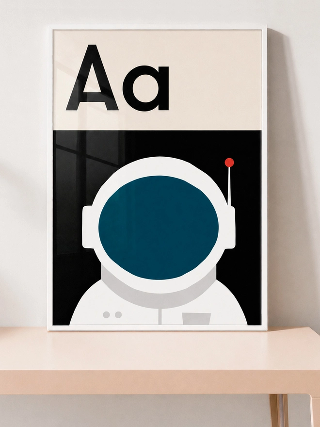 ramed poster with letter 'A' and astronaut illustration on a table against a white wall.