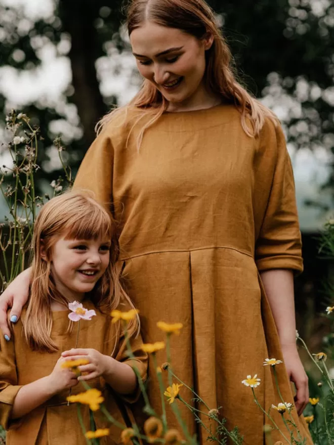 Midi length luxury linen dress in golden colour. Deep pleated skirt and elbow length sleeves. Lifestyle shot outdoors.