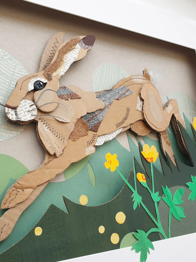 Hare leaping through long grass and buttercups, created using multiple layers of corrugated card, decorated with salvaged fabric and paper. Including buttons and feathers. Finished off with machine and hand embroidery.  