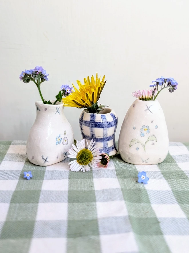 forget me not and blue gingham check miniature vase held on a green gingham cloth with dandelion, daisies and forget me not flowers in and hand painted illustration