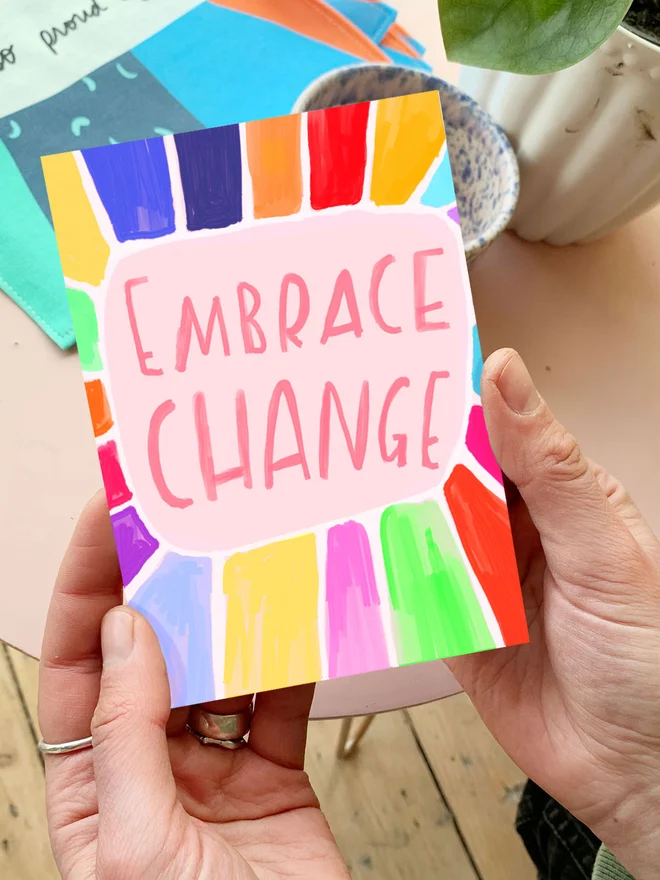 Colourful card reads 'embrace change' in large handwritten font. Hands holding card in lifestyle setting.