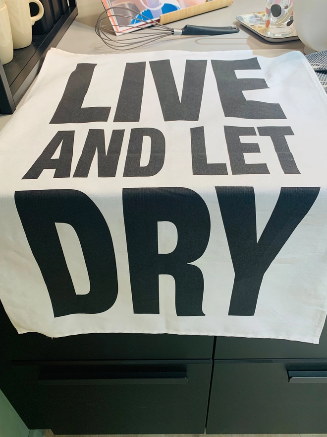 London Drying Live and Let Dry black print on white tea towel laying on a kitchen counter