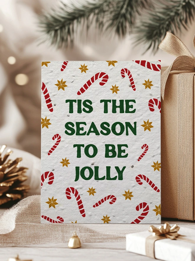 Christmas Card with candy canes and gold star pattern with 'Tis the season to be Jolly' in bold forest green text. The card is surrounded by Kraft paper wrapped presents and pine cones.