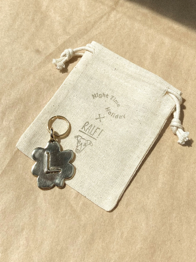 a brass pet tag in the shape of a flower lays in the sun ready to be packed, with a cotton drawstring bag stamped with a whippets face and 'night time holiday X ralf!' 