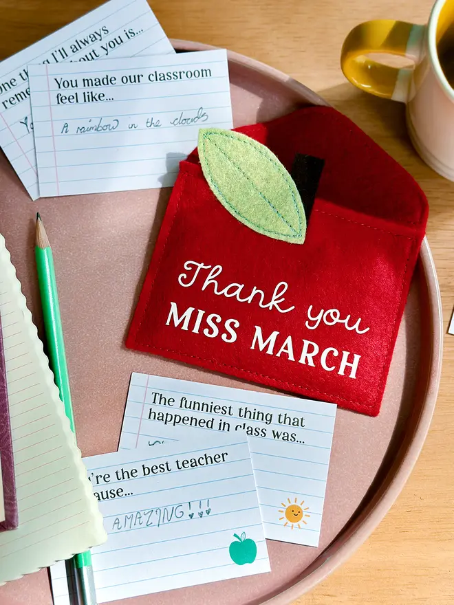 A handmade red felt apple-shaped envelope with "Thank you Miss March" written on the front. The heirloom apple envelope surrounded by completed prompt cards with handwritten messages from a child, displayed on a pink plate and wooden desk. It's a thoughtful personalised gift for teachers from children.