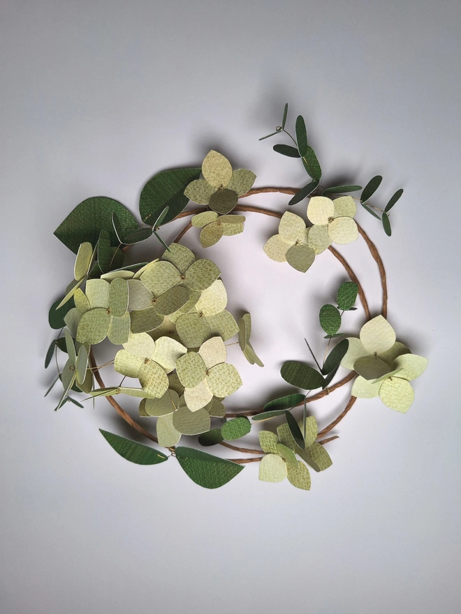 Green hydrangea flower wreath, handmade from paper. There is a cluster of 16 hydrangea blooms on the wreath, which is also adorned with single flowers and two types of dark green leafy stems.