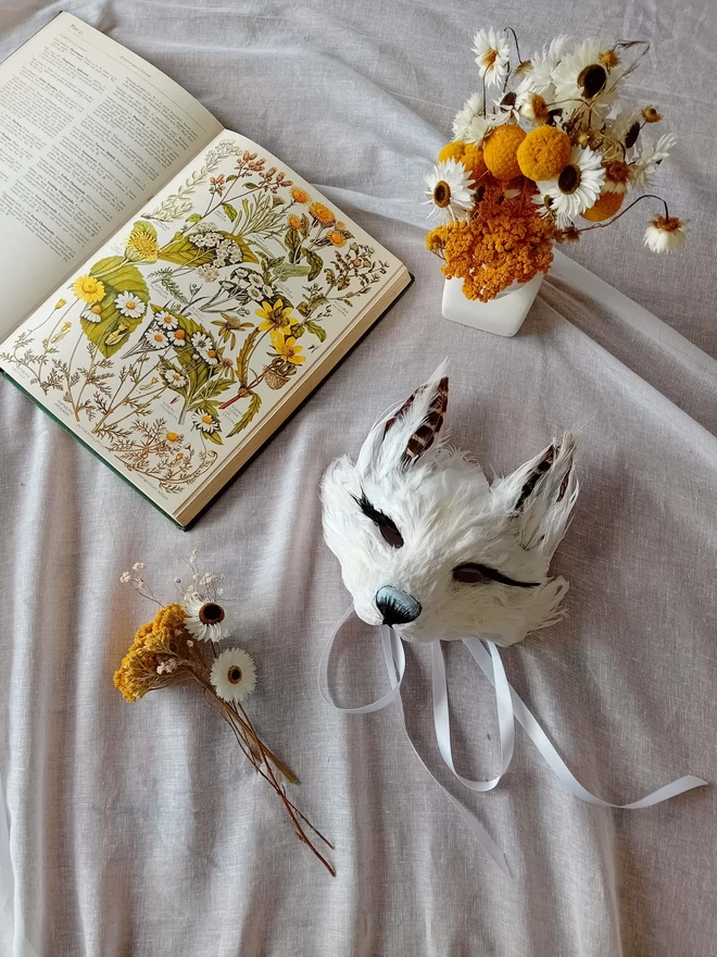 A kid's luxury white fox mask flat lay with yellow and white flowers and a botanical book