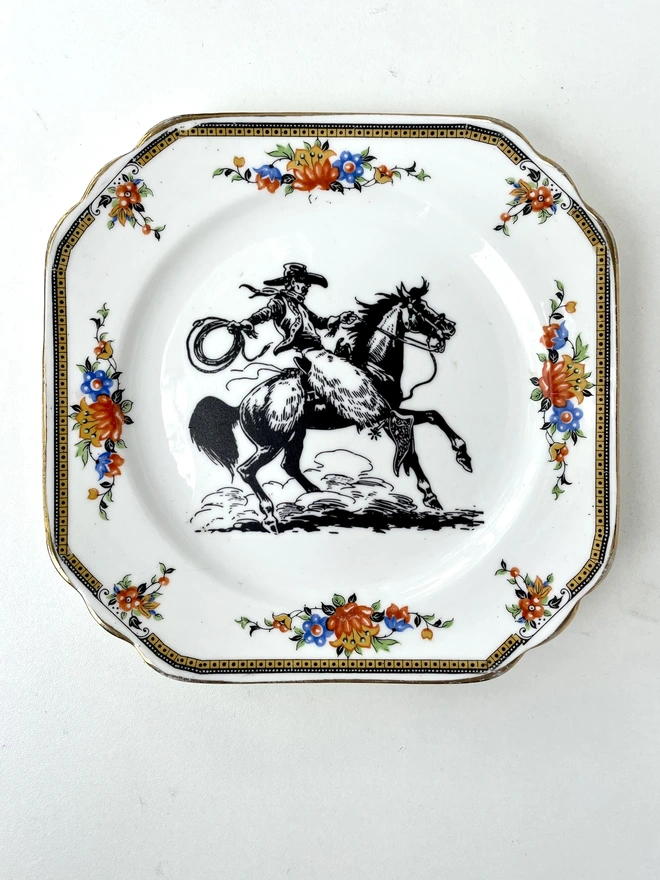 vintage plate hanging on a white wall, it has a decorative border, with a printed vintage illustration of a cowboy in the middle 