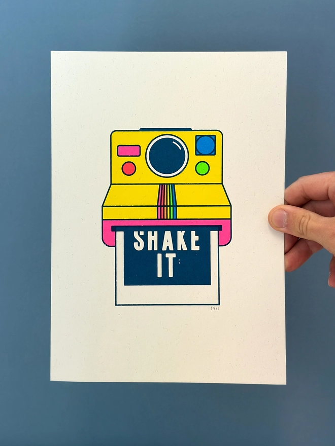 Picture of a hand holding an A4 piece of paper with a screen printed image of a polaroid instant camera in flouro colours. The words shake it can be seen coming out on the instant photo from the camera. 