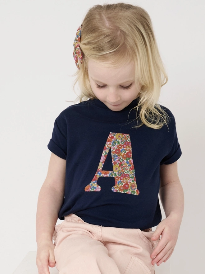 a little girl wearing a personalised kids navy t-shirt with an initial cut from floral liberty floral print on the front