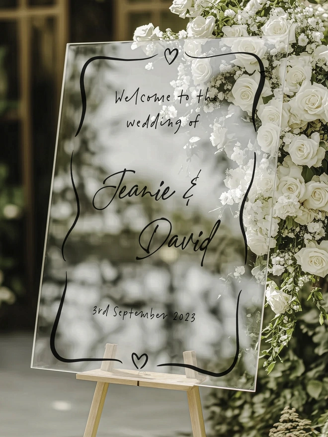 Clear plastic board on a wooden stand with black writing of Welcome to the wedding of Jeanie & DAvid on the 3rd september 2023 surronded by two heart at the top and bottom and some squiggle lines around the words. the board stands in front of a bouqet of white flowers and green foliage