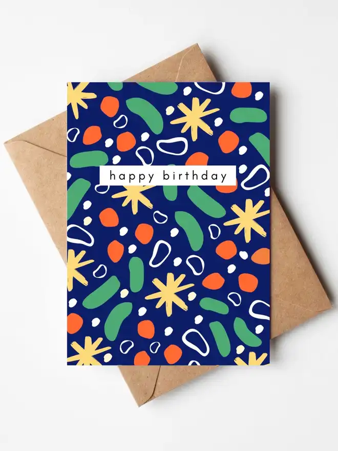 A colourful abstract birthday card featuring a bold blue background with hand-illustrated green, orange, yellow and white shapes, with the words "happy birthday" across the centre. The card is displayed with a kraft envelope.
