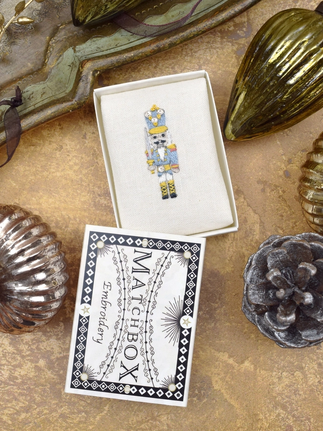 Embroidered nutcracker, inside a Matchbox displayed on a gold background with glass baubles.