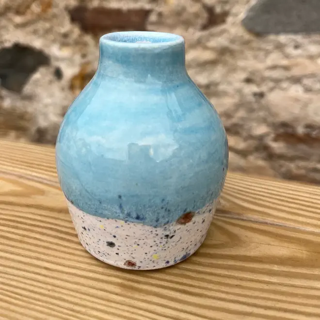 Shoreline Bud Vase, a colourful vase placed on a wooden surface against a brick backdrop. 