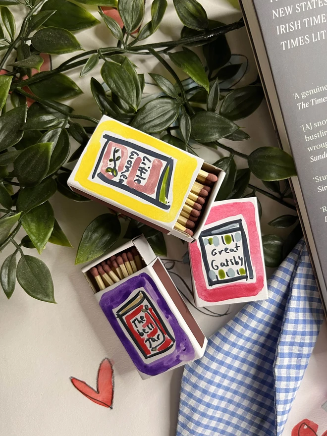 A collection of three hand-painted matchboxes with playful and colorful designs: one features two glasses with the word "Cheers!" written in bold lettering, another displays bright yellow lemons with "Easy Squeezy" written alongside, and the third showcases a red bow with the phrase "Bows & Arrows." Each matchbox is crafted with vibrant illustrations and creative lettering.