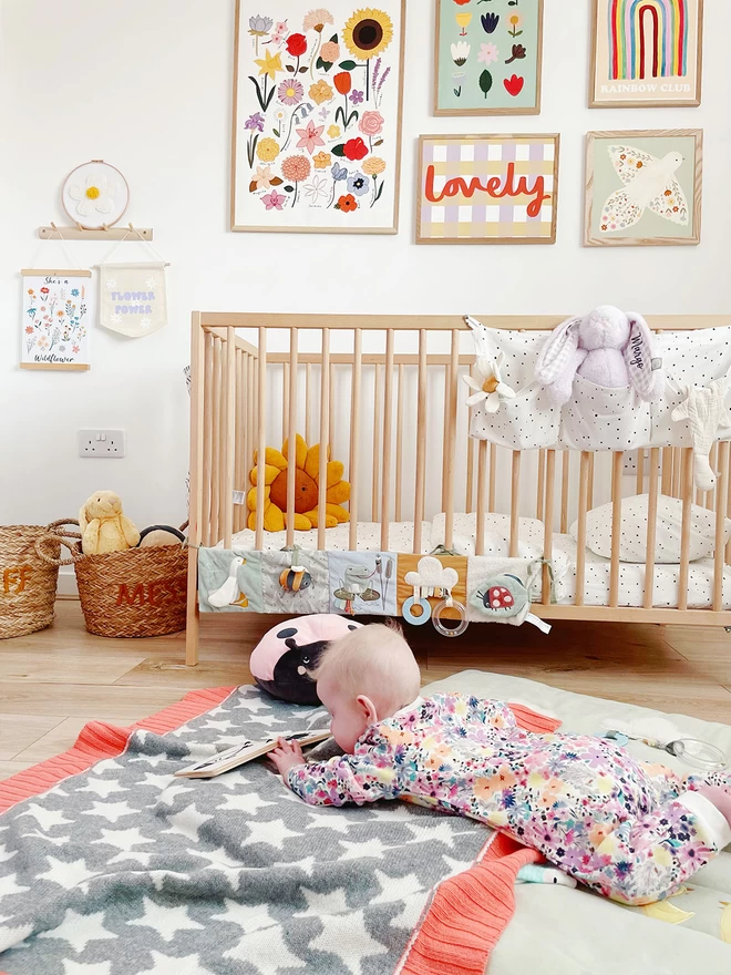 A baby lies on its tummy in a bright and colourful nursery.. The baby is facing away from the camera and is lying on the reverse side of the grey and white baby star blanket with coral pink trim on the floor looking at a book.  