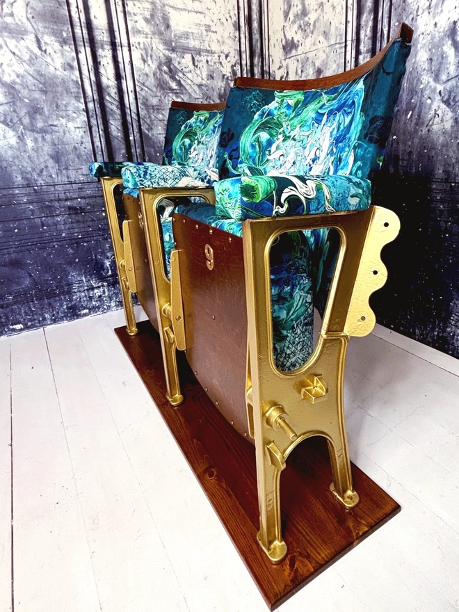 Sideview of a set of vintage cinema seats which have gold legs and blue and green mermaid velvet upholstery.  the seats are sat on a white wooden floor with a blue marbled wall