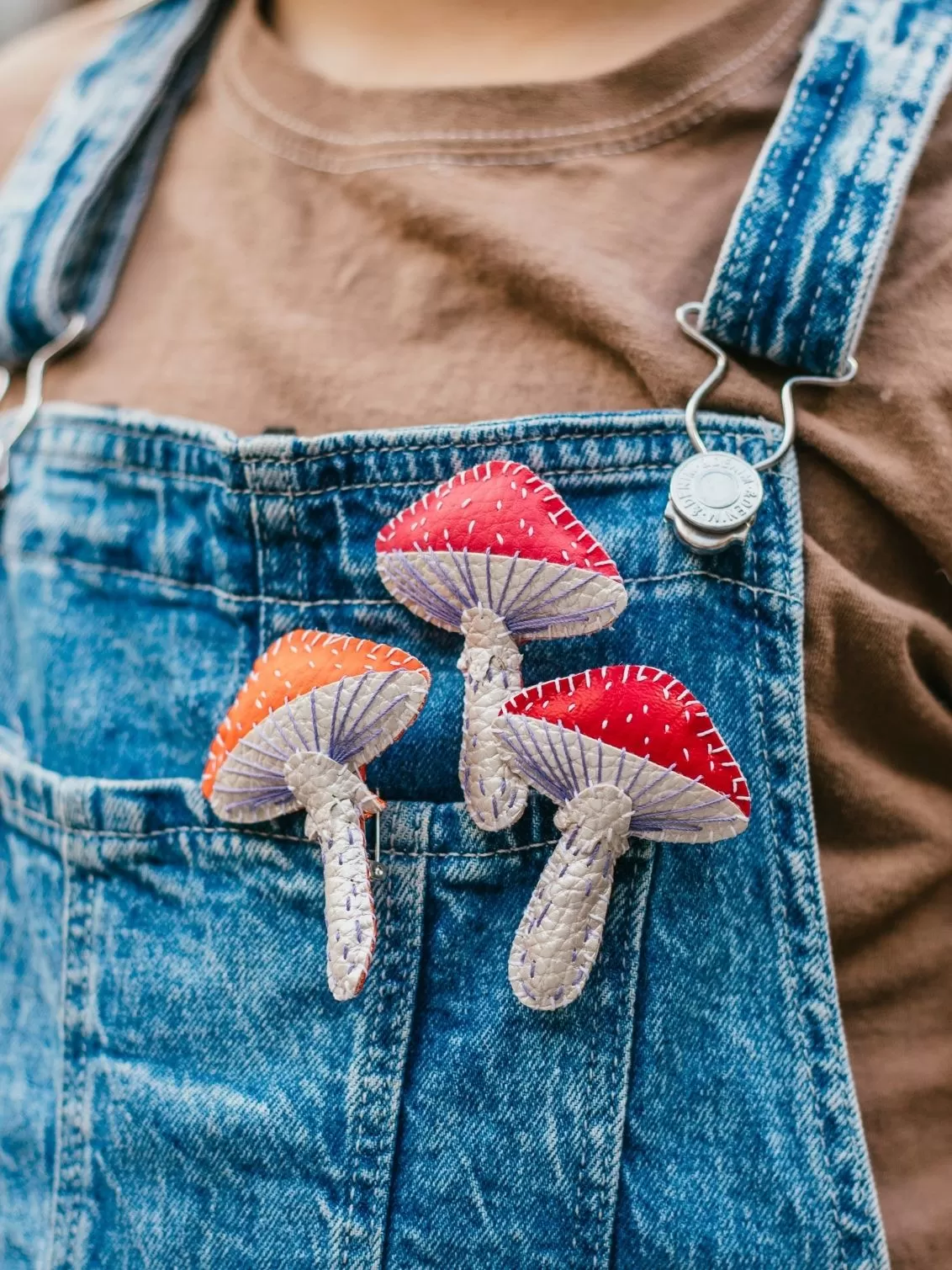 Three hand stitched faux leather toadstool brooches, in red, pink and orange seen on denim dungarees.