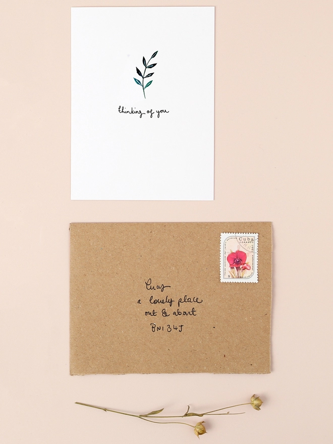 An image of a mini, rectangular white greetings card with an illustration of a sprig of leaves in dark blue ink, with a hand written 'thinking of you' message in black ink seen under the central illustration. The card is placed on top of a brown rectangular kraft envelope.