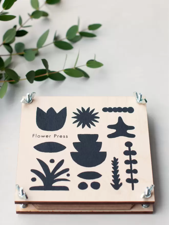 Flower press with black, modern abstract print on the front, sat on a table top with a plant coming in from one corner.