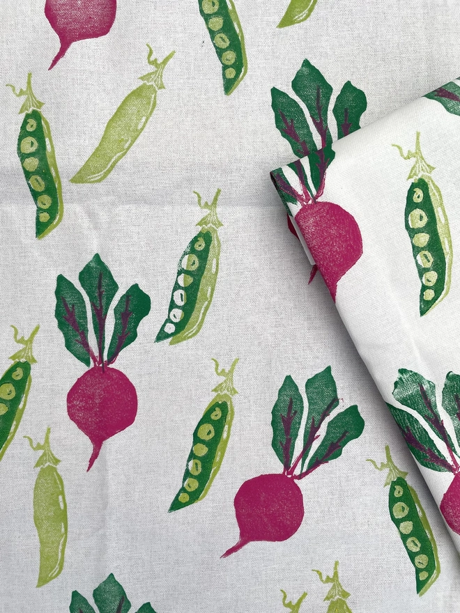 Beetroot And Pea Kitchen Tea Towel. White fabric with a diagonal pattern of green pea pods and beetroots, some open to show bright green peas.