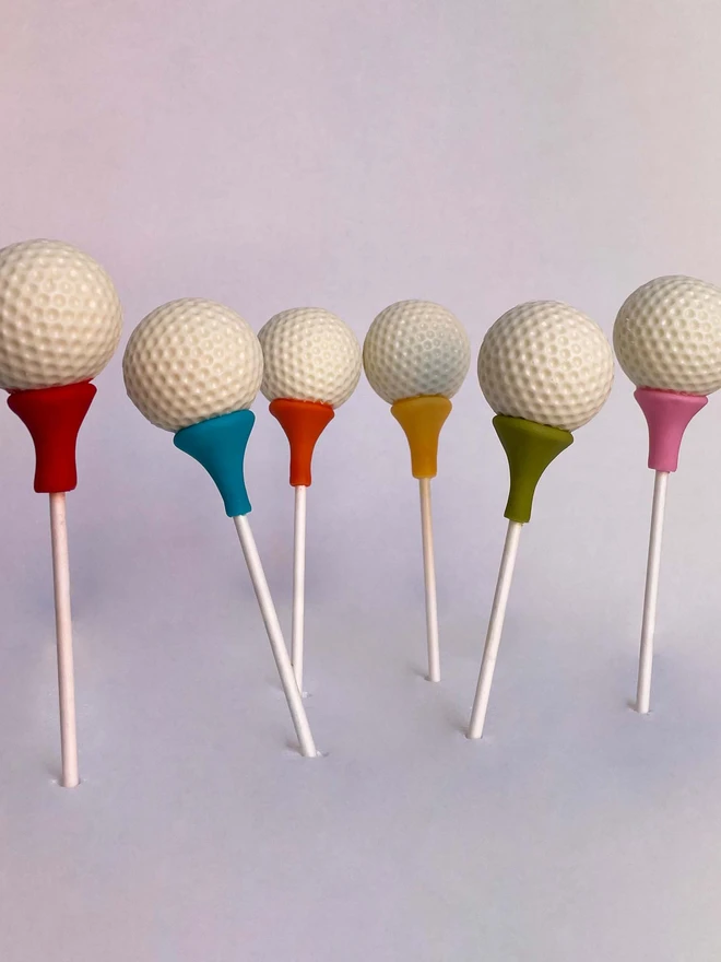 Golf Ball Cake Pops (Pack Of 6, 12, 18, 24), cake pops standing in a row against a off white background. Each tee has it's own colour. 