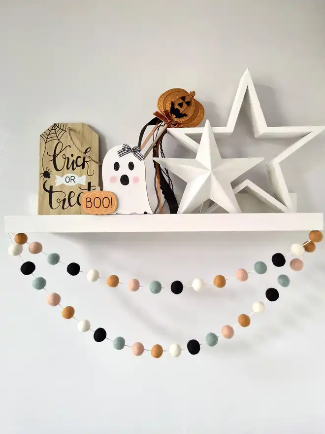 A white shelf filled with Halloween decor. Underneath is hanging a felt ball garland in peach, duck egg, ochre, ivory and black.