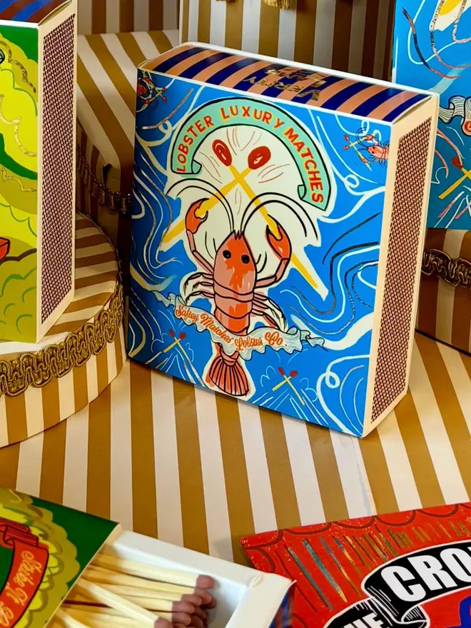Lobster Matchbox, a blue lobster design matchbox sitting on a decorated surface accompanied by other accessories. 