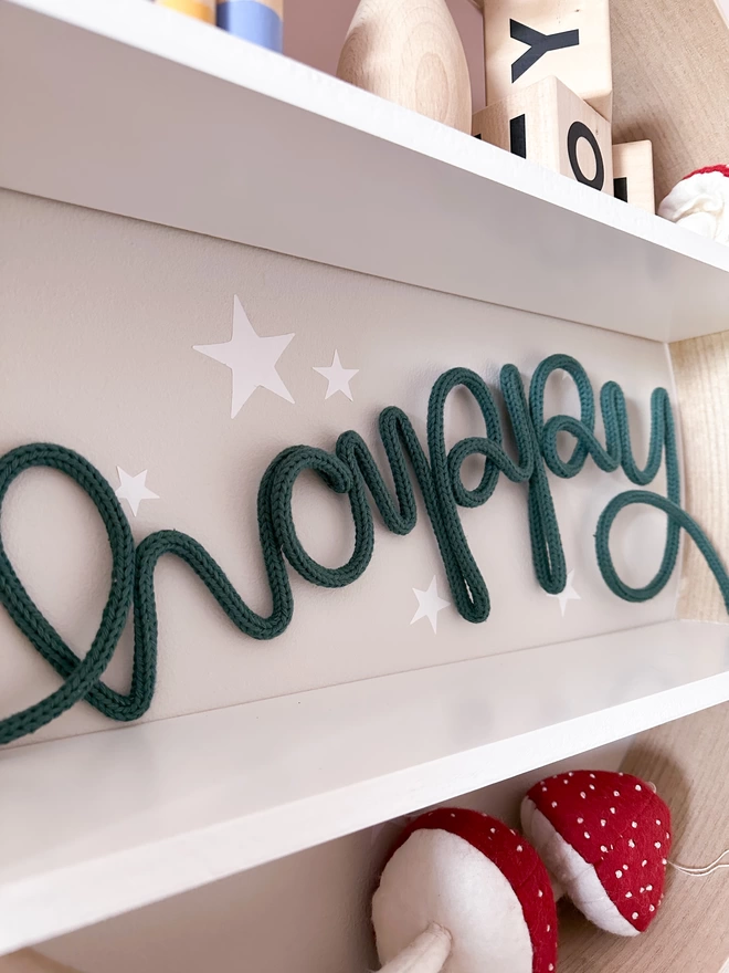 Close up of our knitted 'happy' wall hanging decoration on a white shelf with stars on the wall in the background. 
