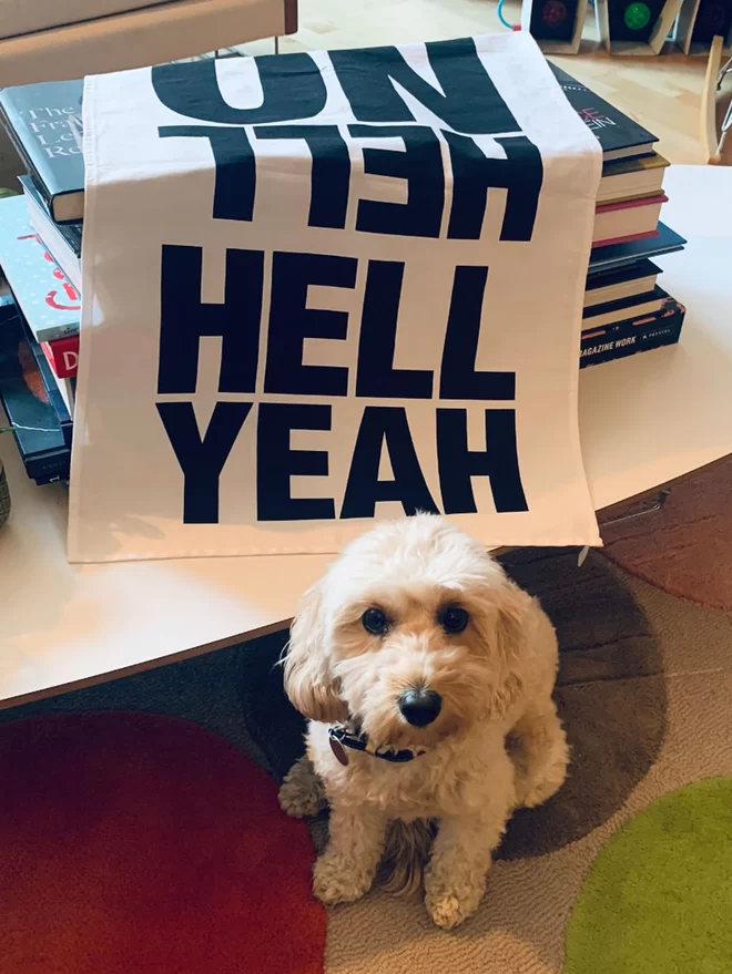 Hell Yeah Hell No screen printed black text on white tea towel laid over pile of books on table with a dog sat in front of 