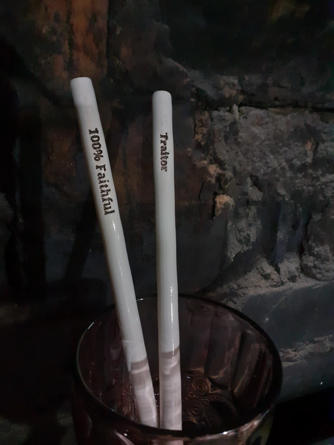 Handmade ceramic drinking straws inspired by The Traitors TV show — one inscribed ‘Traitor’ and the other ‘100% Faithful,’ finished in glossy white glaze with black lettering. The pair stand in a smoky brown glass against a rustic brick wall, softly lit to evoke the show’s moody atmosphere.