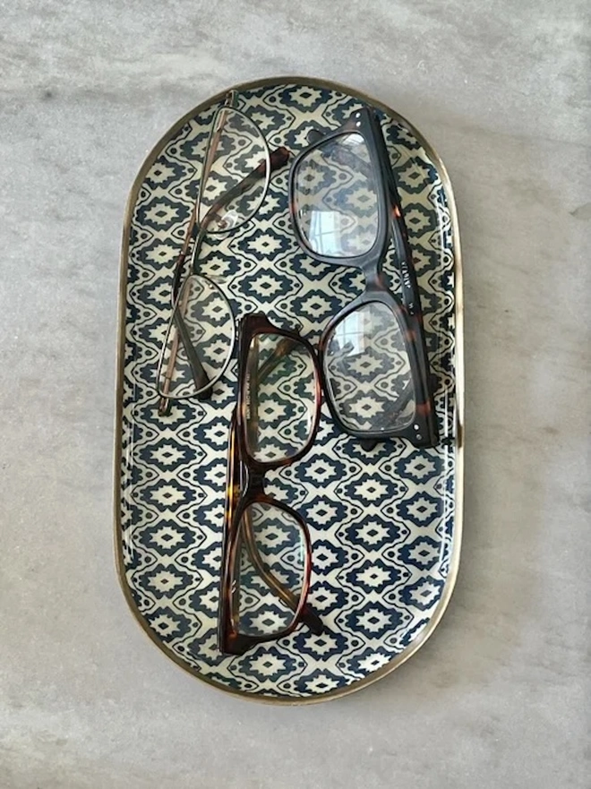 Perfect Enamel Oval Tray. A navy and cream ikat-patterned oval tray holding three pairs of glasses on a grey marble surface. The gold-rimmed catchall tray keeps spectacles, jewellery and everyday essentials neatly together.