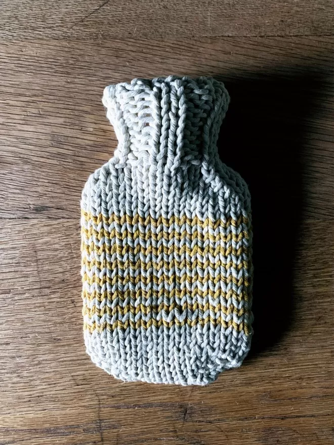 Mini Hot Water Bottle in hand knit string cover with orange stripe on an oak table
