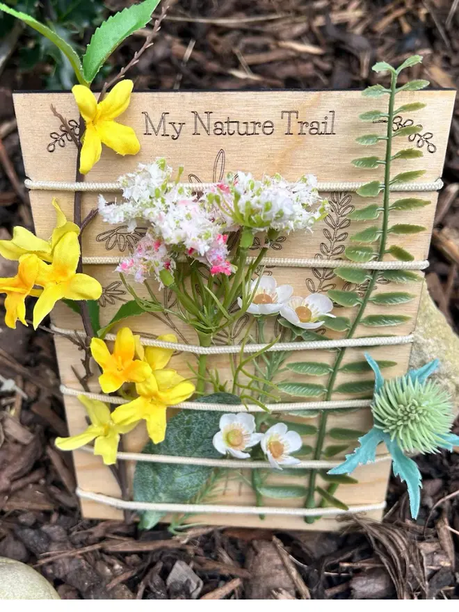 Birch wood nature trail weaving board decorated with artificial wildflowers and leaves, held in place by cream-colored cord, displayed outdoors against mulch and stones.