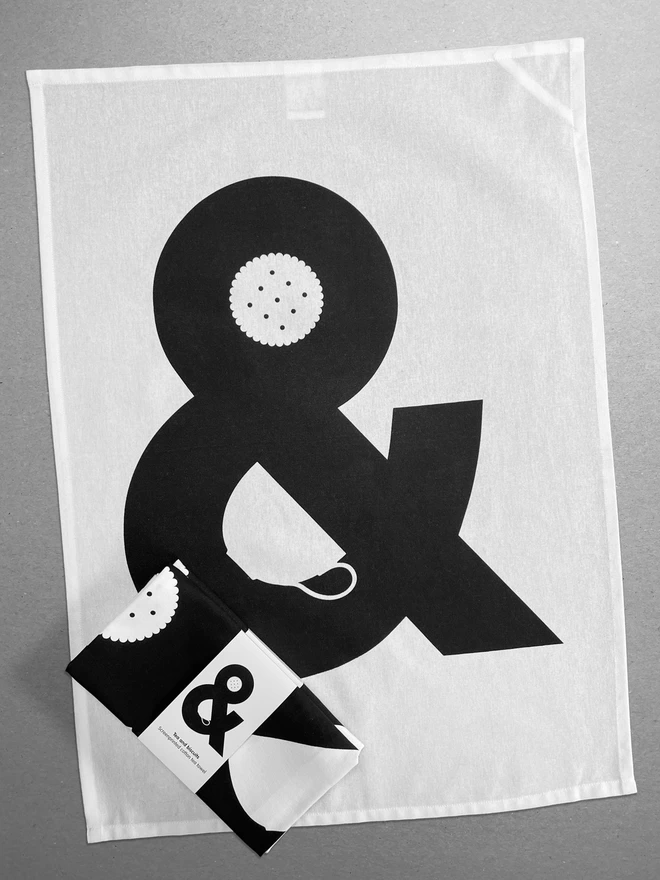 A folded tea towel lies on an opened out tea towel - the design is a giant black ampersand which has a cup and a biscuit hidden in the negative spaces. The folded tea towel has a belly band also showing the complete design.