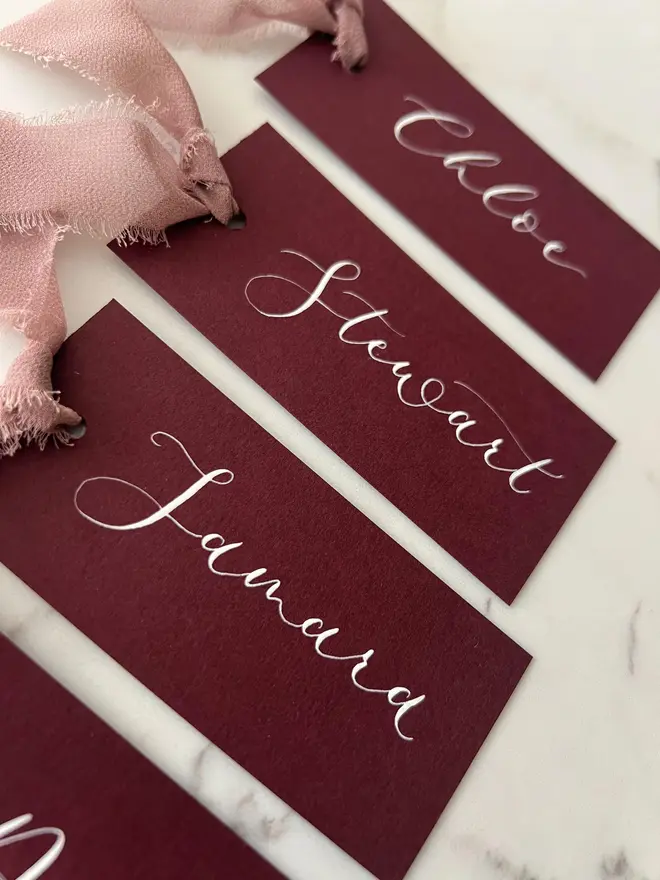Personalised Calligraphy Wedding Burgundy Place Cards, burgundy place cards set together on a wooden table with chiffon plum ribbons. 