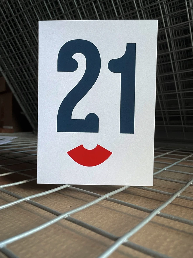The Number 21 screen printed in a dark blue serif typeface. The serifs make the number 21 look like the eyes and nose of a face from a slight angle. Below is a red bracket on its side. This looks like the lips of the face. The image has an Art Deco feel to it. The portrait card is stood straight on, on a drying rack.