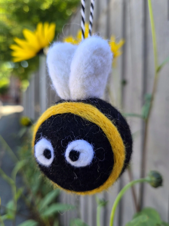Black and yellow felt bee decoration, hanging with a black and white Bakers twine in front of some flowers.