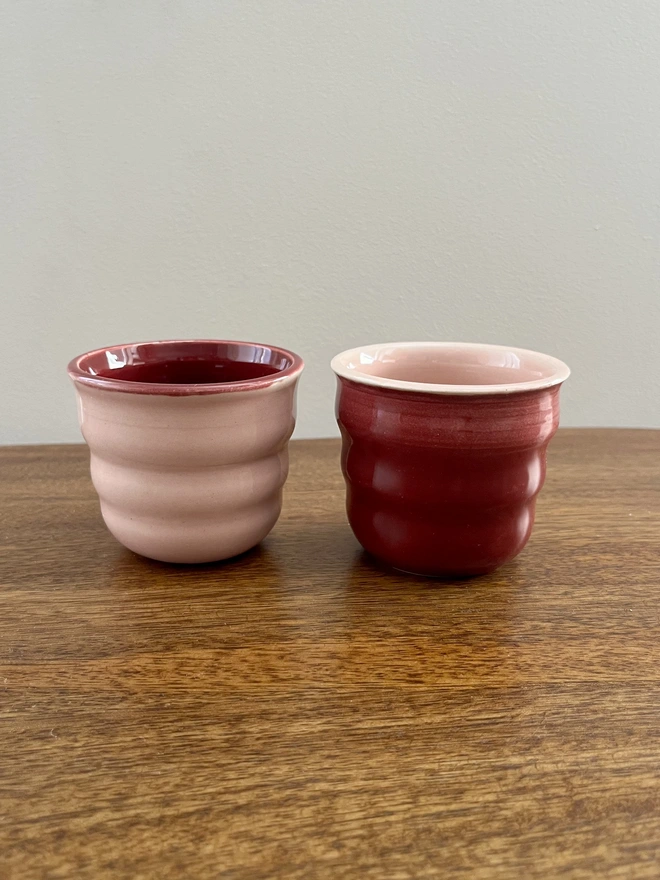 a pair of ceramic tumblers with a wavy sides on a wooden table. the pair is glazed in reverse colours. one is blush pink on the outside and sangria burgundy on the inside and the other is the opposite with the sangria burgundy on the outside and blush pink on the inside.  