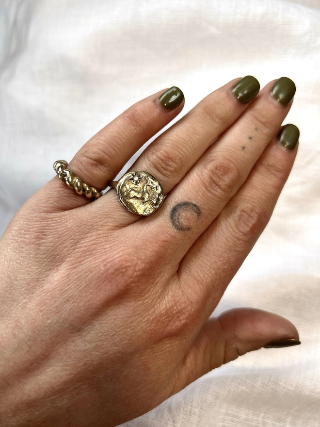 Image of a hand wearing a gold toned brass ring with a horse and star on it, and another twisted metal style gold toned brass ring. The back ground of the image is white linen 