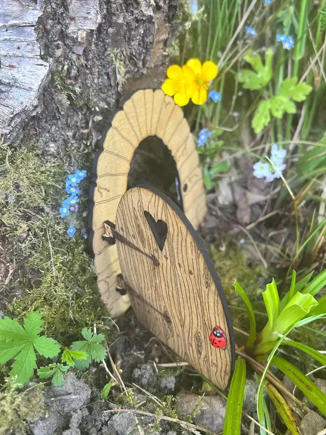 Wooden fairy door for kids with engraved woodgrain detail, heart-shaped window, and decorative hinges, displayed at the base of a tree surrounded by moss and wildflowers.